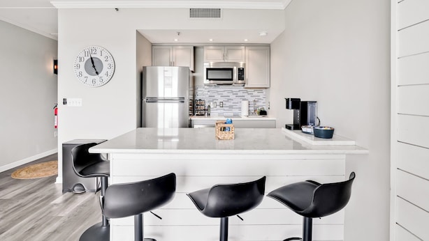 A modern kitchen with a central island, stainless steel appliances, and a clock on the wall. - Battleship Sunset on Water Street! River View! Free parking!! (Wilmington)
