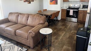 Een woonkamer met een bruine leren bank, een eettafel met stoelen en een keuken met houten kastjes en apparatuur.