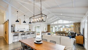 A spacious open-plan living area with a wooden dining table, multiple chairs, a sofa, and a kitchen area with white cabinets and a large window. - Modern Deluxe - Havelock North Holiday Home (Havelock North)