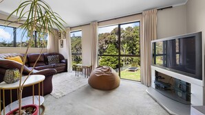 Un salon avec un canapé en cuir marron, une table basse en verre, un fauteuil poire beige, un téléviseur à écran plat posé sur un meuble, et une grande fenêtre donnant sur des arbres et une pelouse.