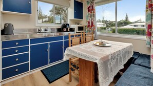 Cocina compacta con armarios azules, microondas y cafetera. Hay una mesa de comedor con mantel de encaje y un banco.