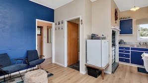 Un espacio habitable compacto con kitchenette, una zona de comedor con pared azul y una sala de estar con sofá azul y reposapiés redondo.