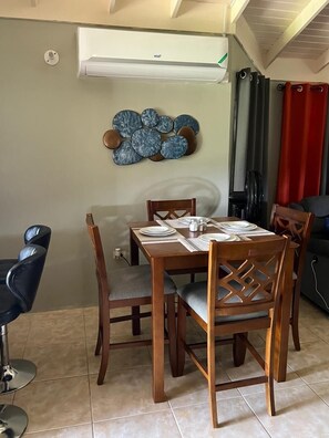 A dining area with a wooden table set for four, a modern air conditioning unit above, and a decorative wall piece. - Reddie Vacation Home at Stonebrook Vista Trelawny (Florence Hall Village)