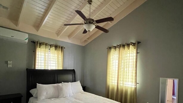 A bedroom with a bed, ceiling fan, and large windows with curtains. - Reddie Vacation Home at Stonebrook Vista Trelawny (Florence Hall Village)