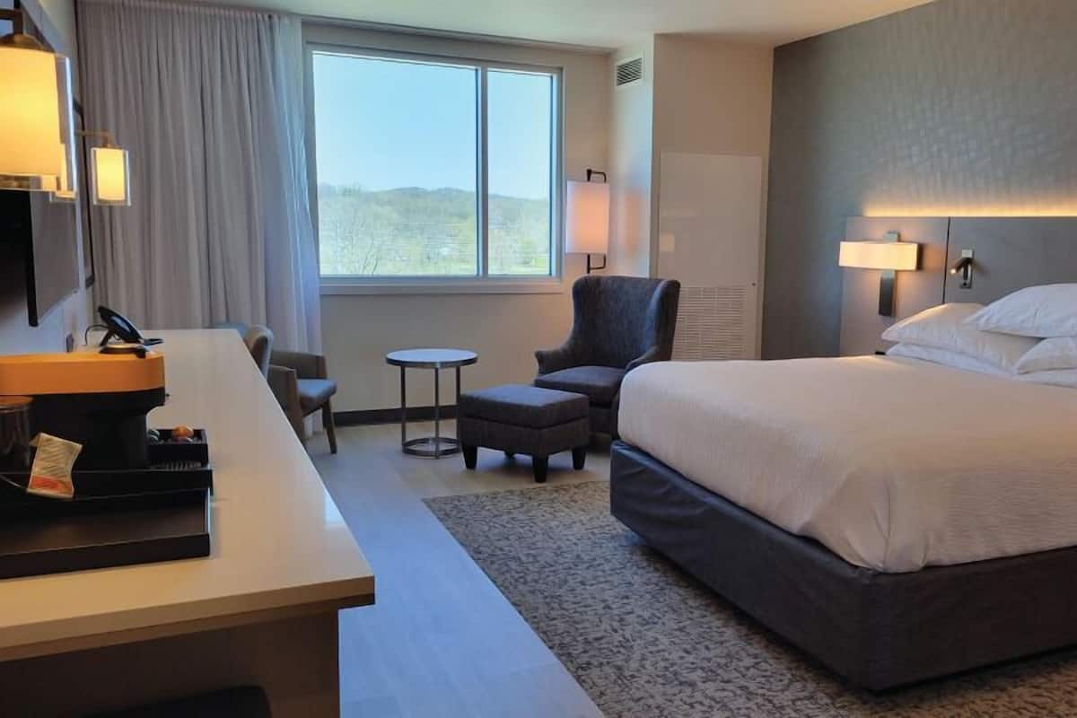 A hotel room with a large bed, a desk, a chair, a small table, and a window with a view of the outdoors.
