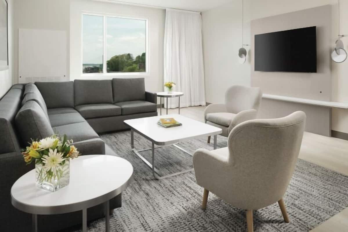 A modern living room with a grey sectional sofa, a round coffee table, a beige armchair, and a flat-screen TV mounted on the wall.