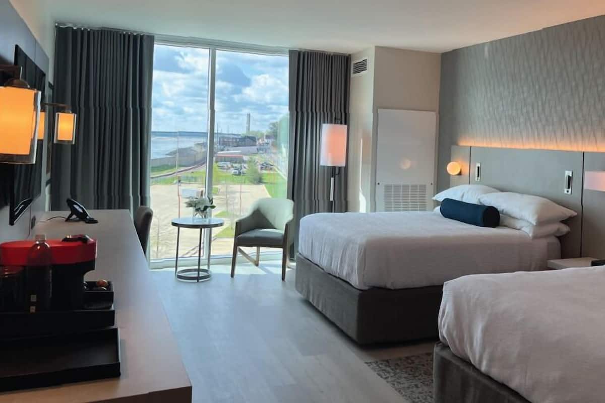 A hotel room with two beds, a desk, a chair, and a view of the city.
