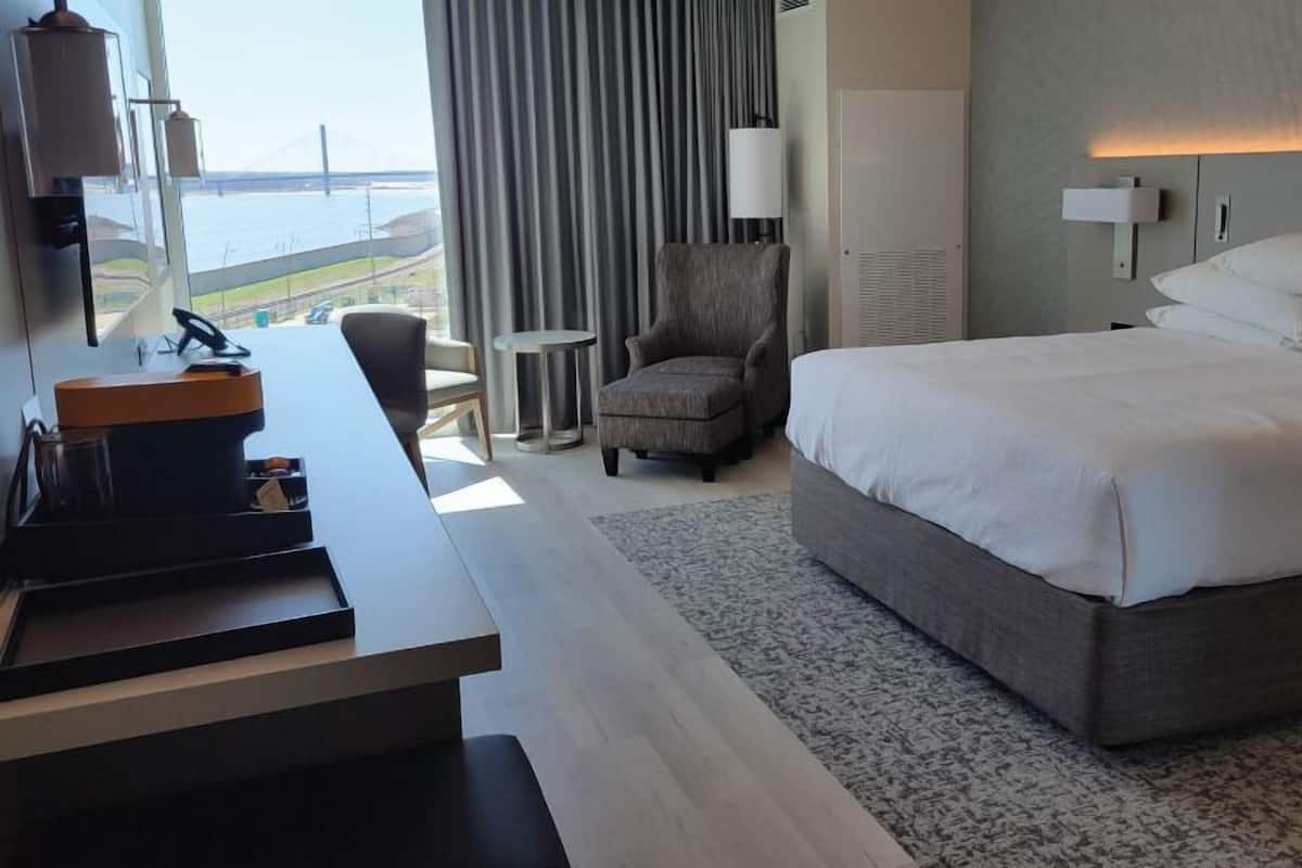 A hotel room with a large bed, a desk with a coffee machine, a chair, and a view of the sea.
