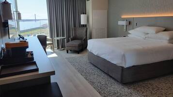 A hotel room with a large bed, a desk with a coffee machine, a chair, and a view of the sea.
