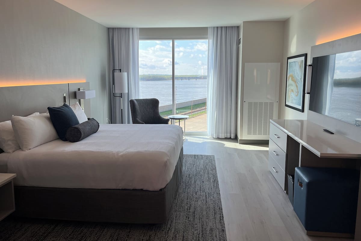 A modern hotel room with a large bed, a desk, a chair, and a TV. The room has a view of the water and a balcony.