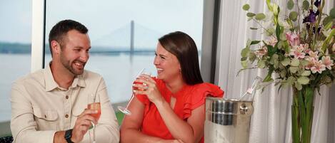 A couple seated at a table with a view of a bridge, a vase of flowers, and a champagne bucket.