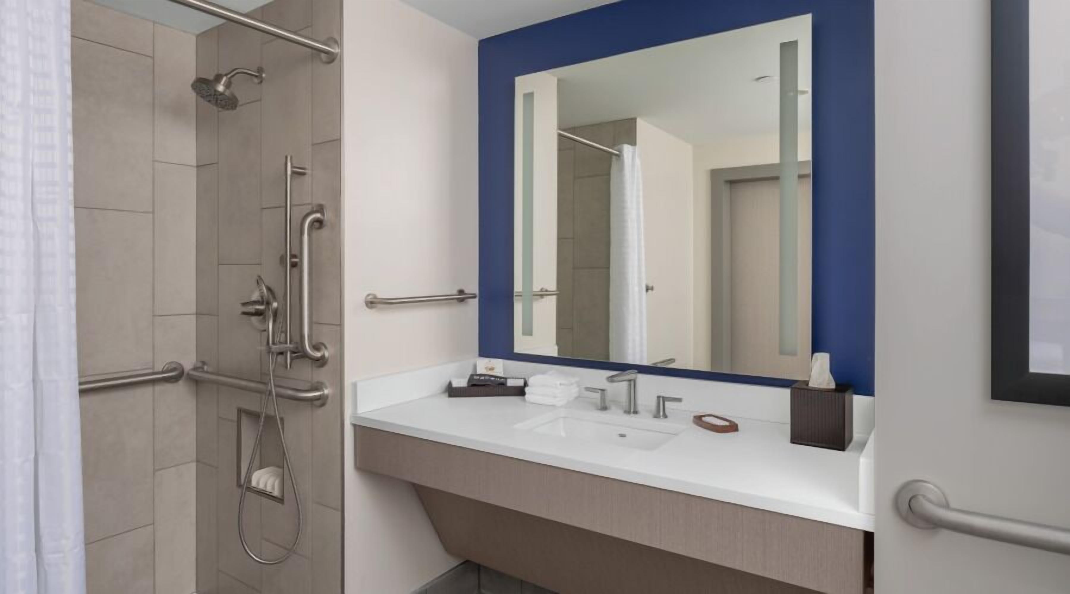 Un baño moderno con ducha a nivel, barras de apoyo, un espejo grande y un tocador blanco con dispensador de toallas.