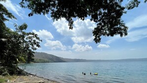A beach with a rocky shoreline, calm water, and a distant hilly landscape under a partly cloudy sky.