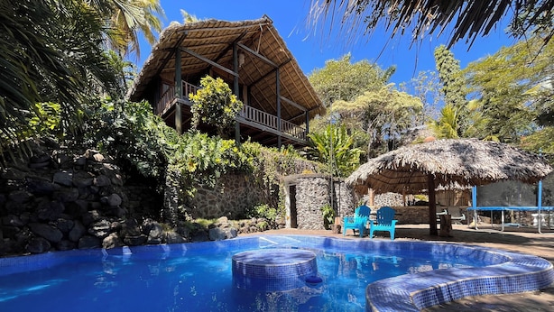 A swimming pool with blue tiles, surrounded by a stone wall and tropical vegetation.