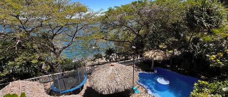 Zona de piscina con trampolín, sombrillas de paja y vista al mar.