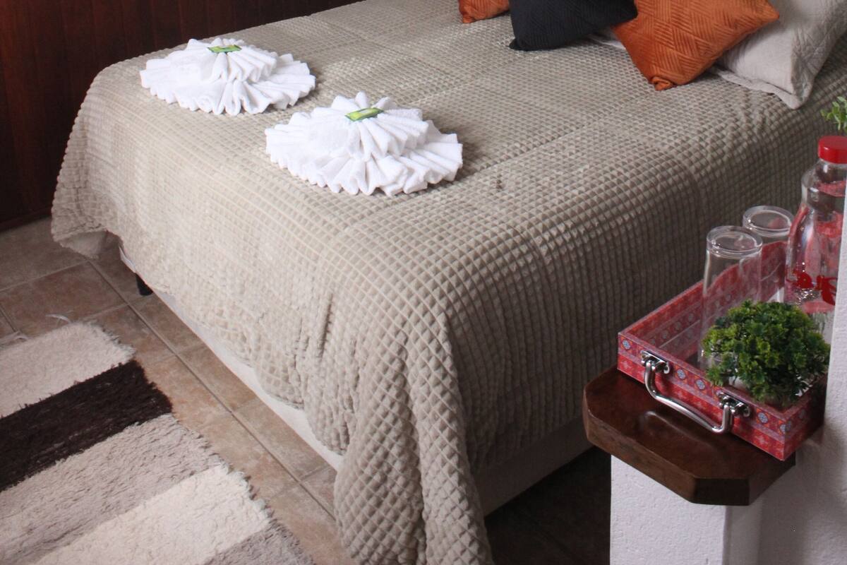 Een net opgemaakt bed met een beige sprei, twee oranje kussens en een zwart kussen. Boven het bed hangen twee kunstwerken aan de muur.