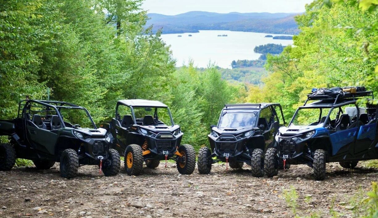 Three off-road vehicles parked on a dirt path surrounded by trees, with a lake and hills in the background.