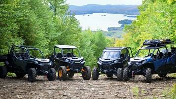 Three off-road vehicles parked on a dirt path surrounded by trees, with a lake and hills in the background.