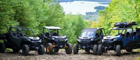 Three off-road vehicles parked on a dirt path surrounded by trees, with a lake and hills in the background.