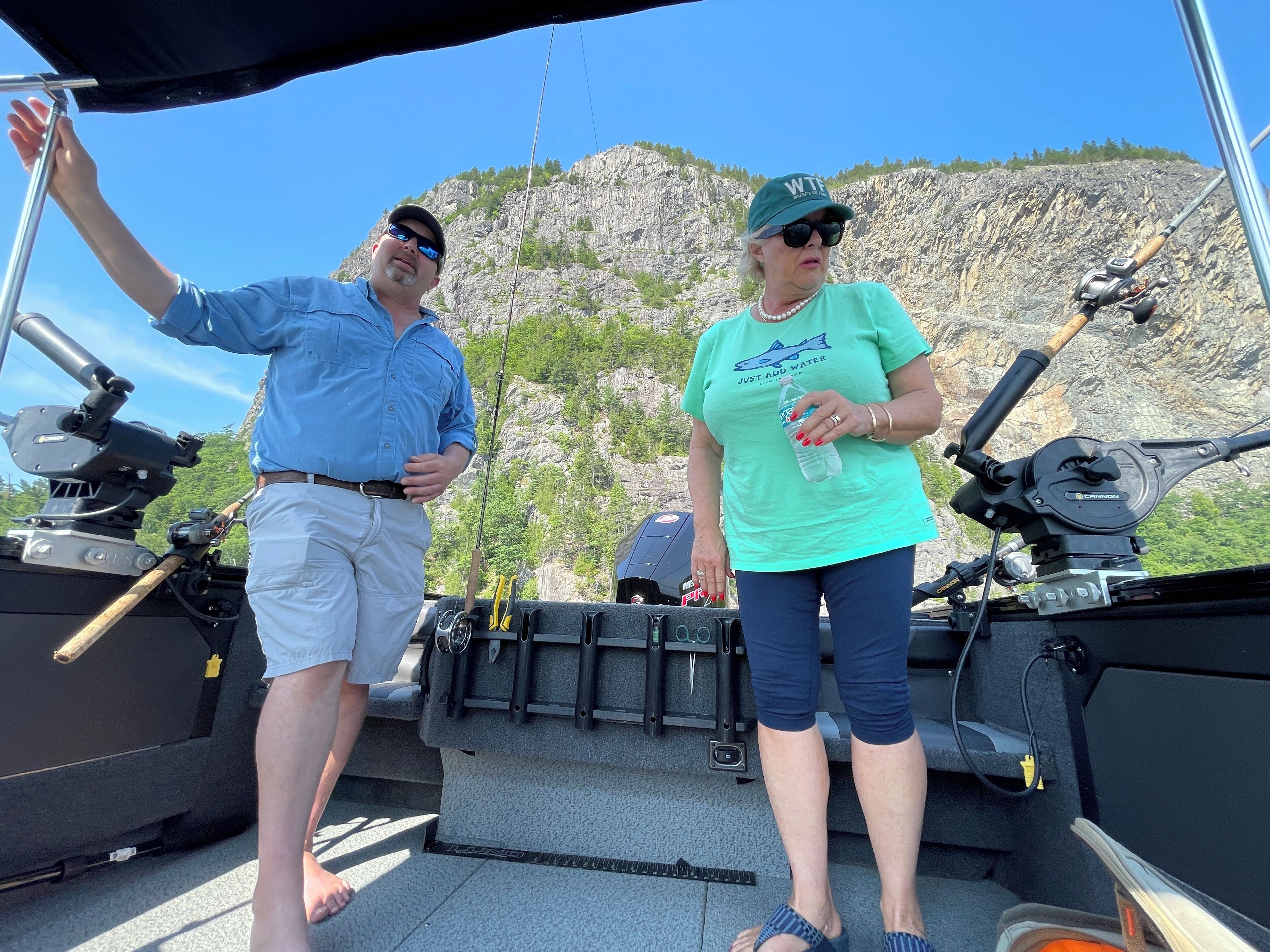 Two people on a boat with fishing equipment, a rocky cliff in the background, and a clear sky.