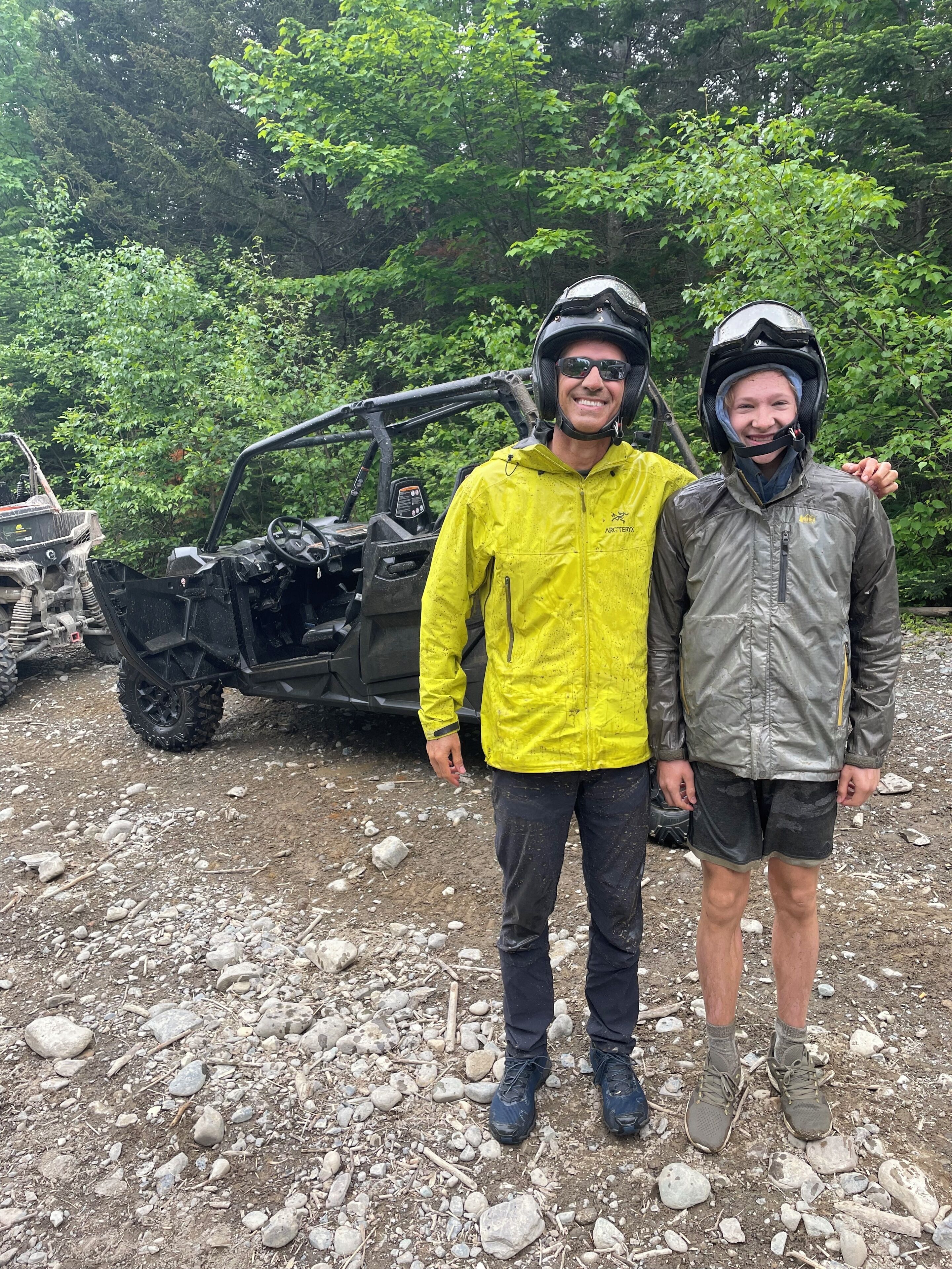 Deux personnes se tiennent devant un véhicule, avec un terrain rocailleux et des arbres en arrière-plan.