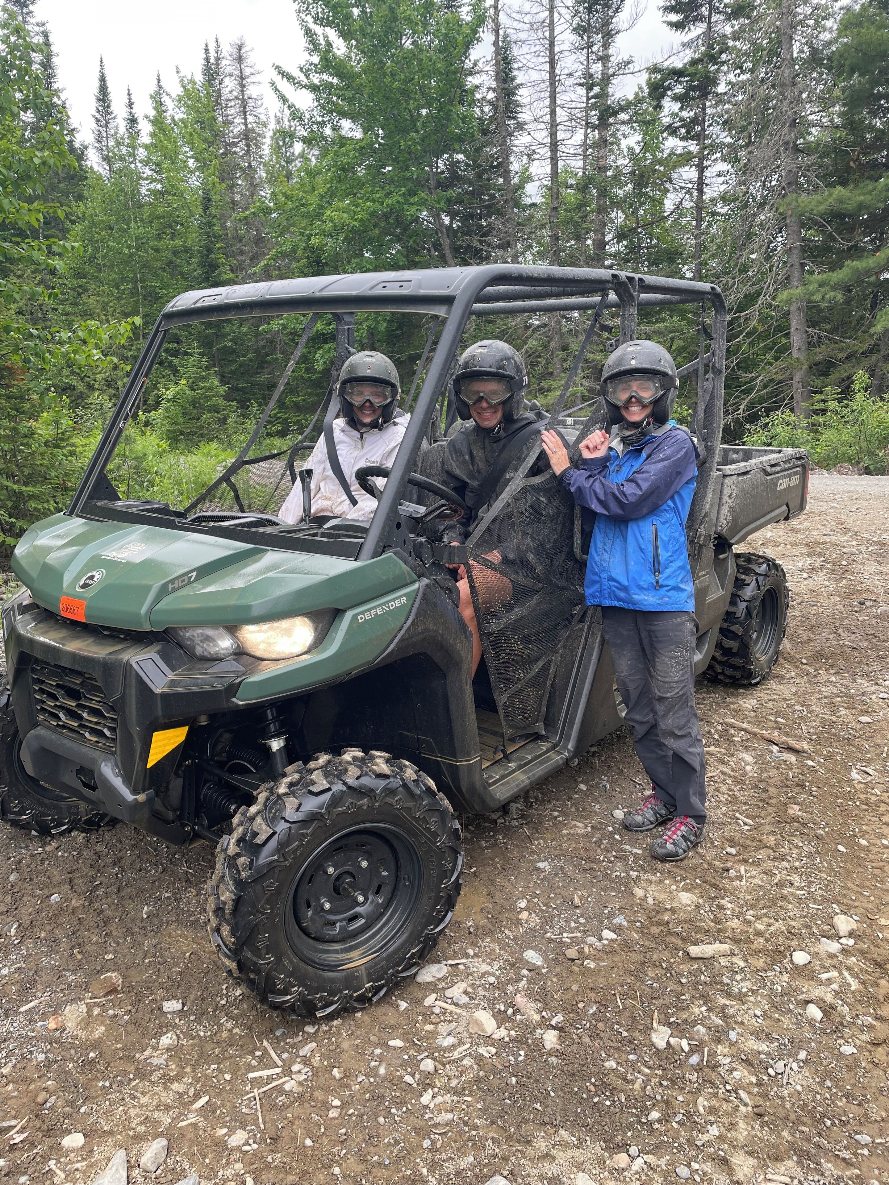 A green utility vehicle with a driver and a passenger, both wearing helmets and protective gear, parked on a gravel surface with trees in the background.