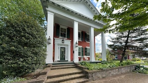 Un bâtiment à deux étages, doté d’une façade blanche et de détails en briques rouges, avec une entrée centrale accessible par des marches et une porte verte.