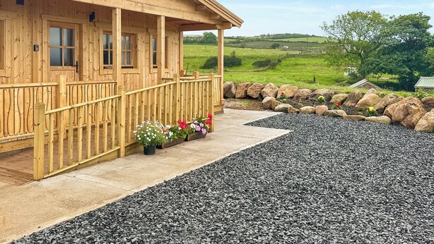 A wooden cabin with a porch, a gravel driveway, and a view of a green landscape. - 2 bedroom accommodation in Seascale, near Gosforth and Wasdale (Seascale, near Gosforth and Wasdale)