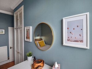 A room with a blue wall, a white door, and a white table. There are framed pictures on the wall, a round mirror, and a wooden bowl on the table.