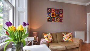 Uma sala de estar com um sofá bege, almofadas coloridas, um vaso com tulipas e um quadro na parede.