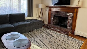A living room with a sectional sofa, a flat-screen TV, a brick fireplace, and a patterned rug. - Charming Cape Cod house in Dennis Massachusetts. Pets Friendly! (Dennis)