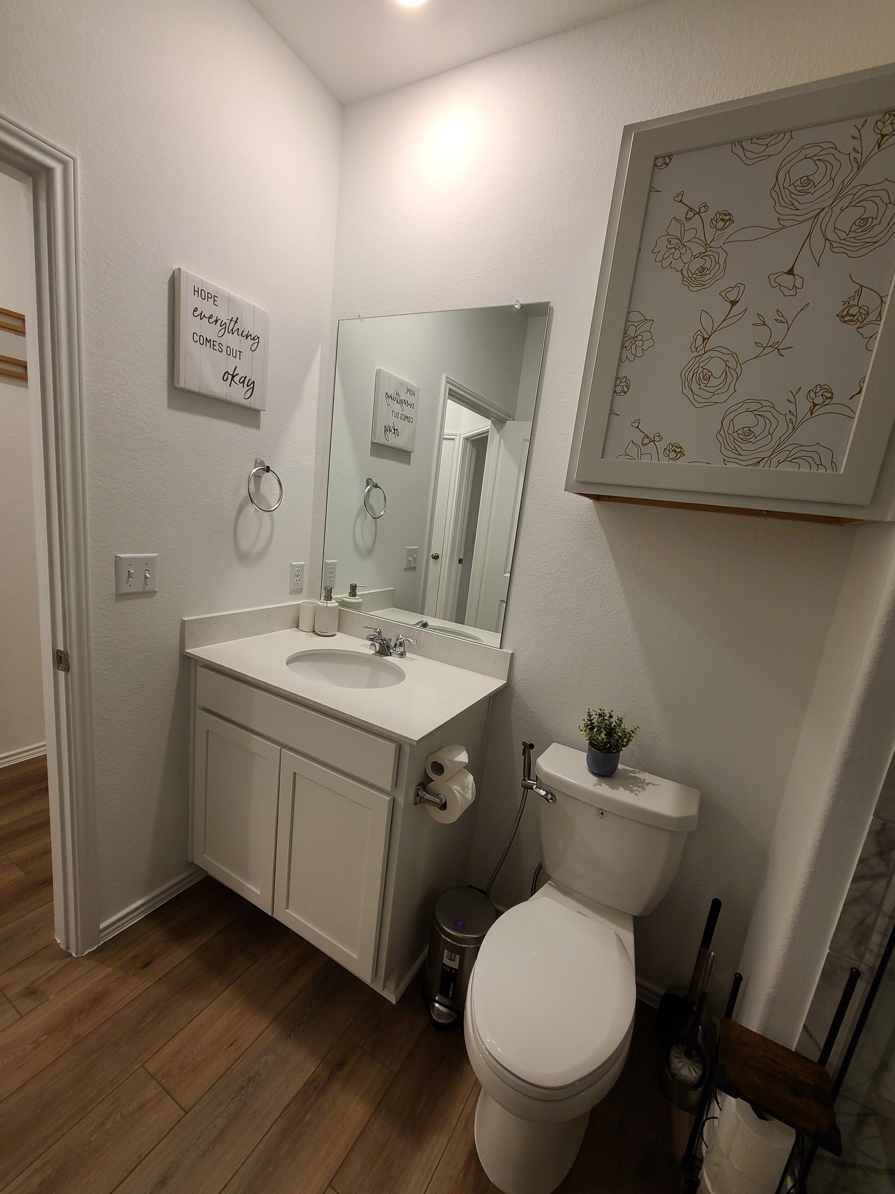 Un baño con lavamanos blanco, espejo, inodoro y una obra de arte floral enmarcada en la pared.