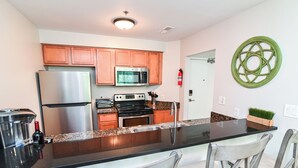 A modern kitchen with wooden cabinets, stainless steel appliances, a black countertop, and bar-style seating. - Bright 2BR Retreat w/ Pool – 1.5 Blocks to Oceanfront & Boardwalk (Virginia Beach)
