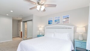 A bedroom with a large bed, two bedside tables with lamps, a ceiling fan, and three wall-mounted pictures. - Bright 2BR Retreat w/ Pool – 1.5 Blocks to Oceanfront & Boardwalk (Virginia Beach)