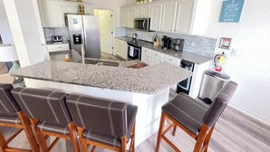 A modern kitchen with white cabinets, granite countertops, a stainless steel refrigerator, and a bar area with leather barstools. - 4BR Villa Bright and Spacious w/ Pool (Davenport)