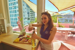 Una mujer sentada a una mesa con una botella de alcohol, una bandeja de comida y una ventana que ofrece vistas a edificios.