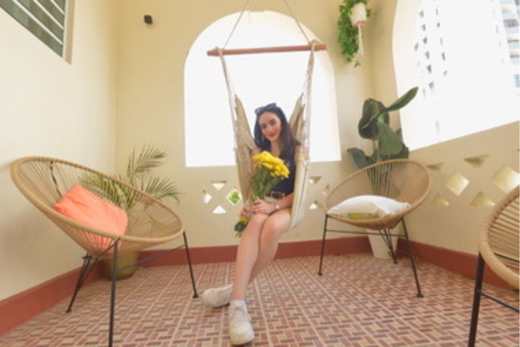 Une pièce avec un sol à motifs, un fauteuil suspendu orné d’une fleur jaune et deux fauteuils en osier avec des coussins.