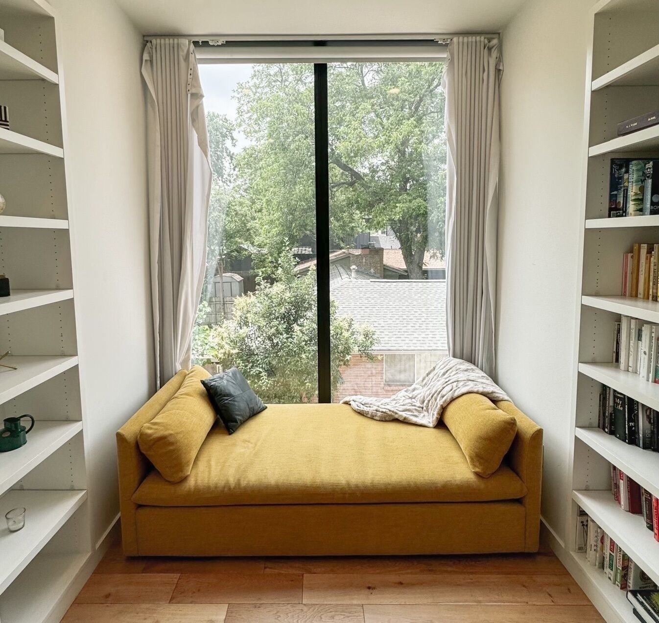 Une pièce avec un canapé jaune, une grande fenêtre avec des rideaux et des étagères à livres de chaque côté.