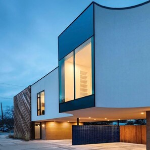 A modern two-story building with a large glass window on the second floor, a curved facade, and a brick wall on the left side. - The Austin Oasis: Modern, spacious, and close to everything (Austin)