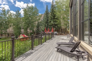 A wooden deck with lounge chairs, a barbecue grill, and a view of a garden with trees and a building in the background. - Pines #2071 By Summit County Mountain Retreats (Keystone)