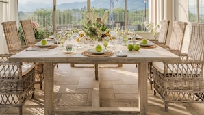 A dining area with a table set for six, surrounded by wicker chairs, under a glass roof with a view of mountains. - Villa Sunkiss Winery (Montecarlo)