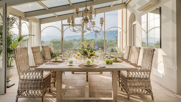 A dining area with a glass ceiling, wicker chairs, a central table set for a meal, and a view of mountains. - Villa Sunkiss Winery (Montecarlo)