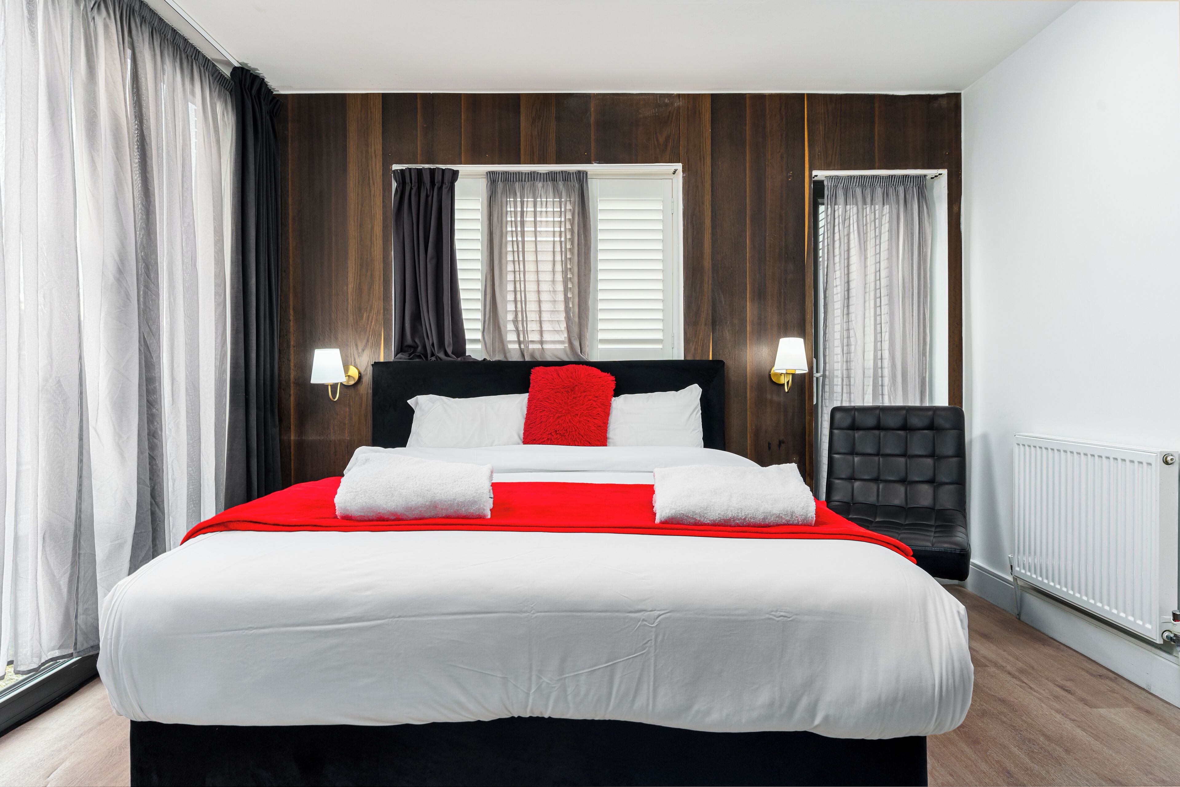 Een netjes opgemaakt bed met wit en rood beddengoed, een zwart hoofdbord en twee bedlampjes.