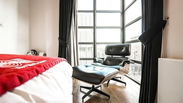 A hotel room with a large bed, a black leather chair, and a view through a large window. - The Whitechapel (London)