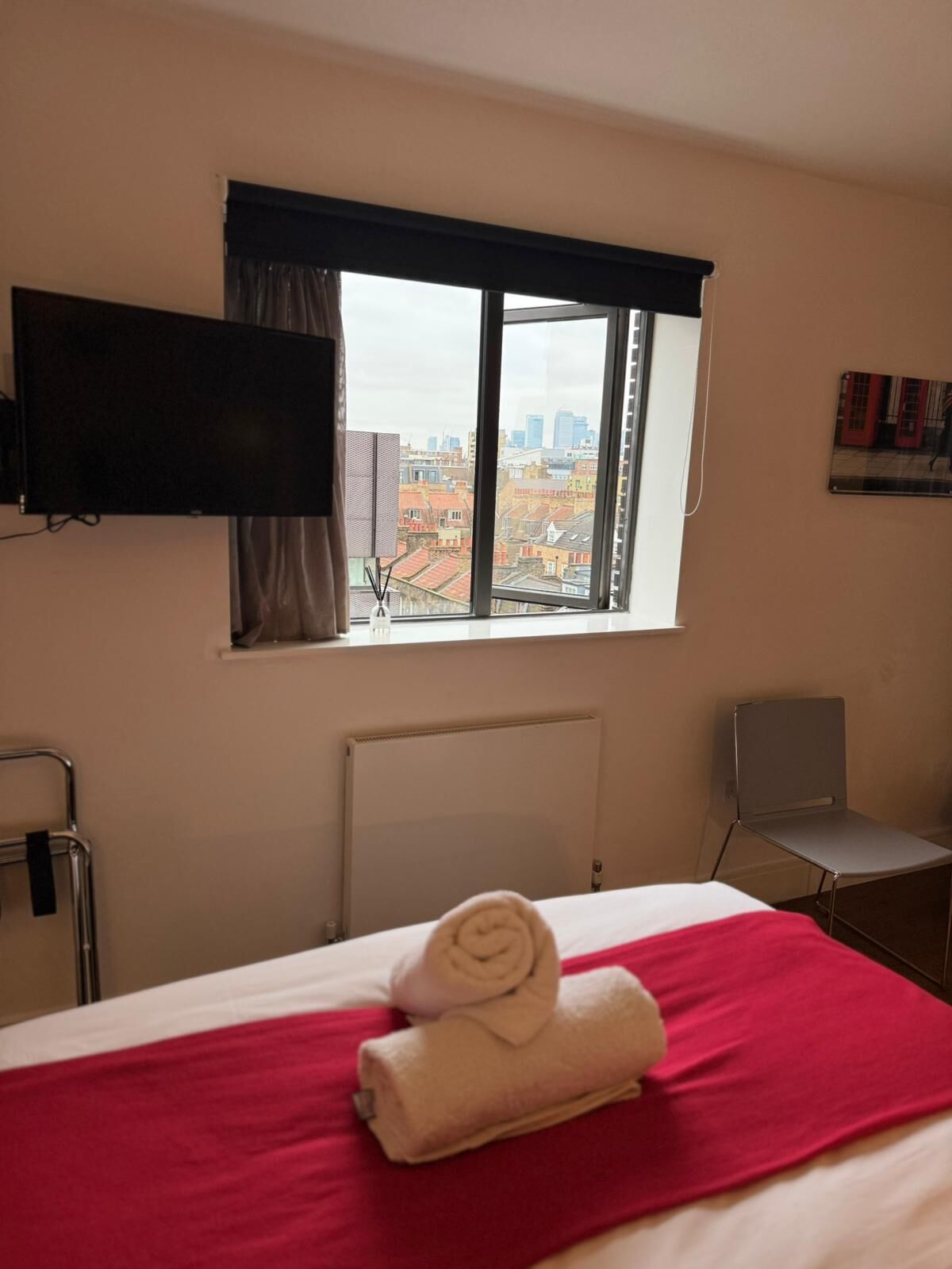 A hotel room with a bed, a window with a city view, a chair, and a TV mounted on the wall.