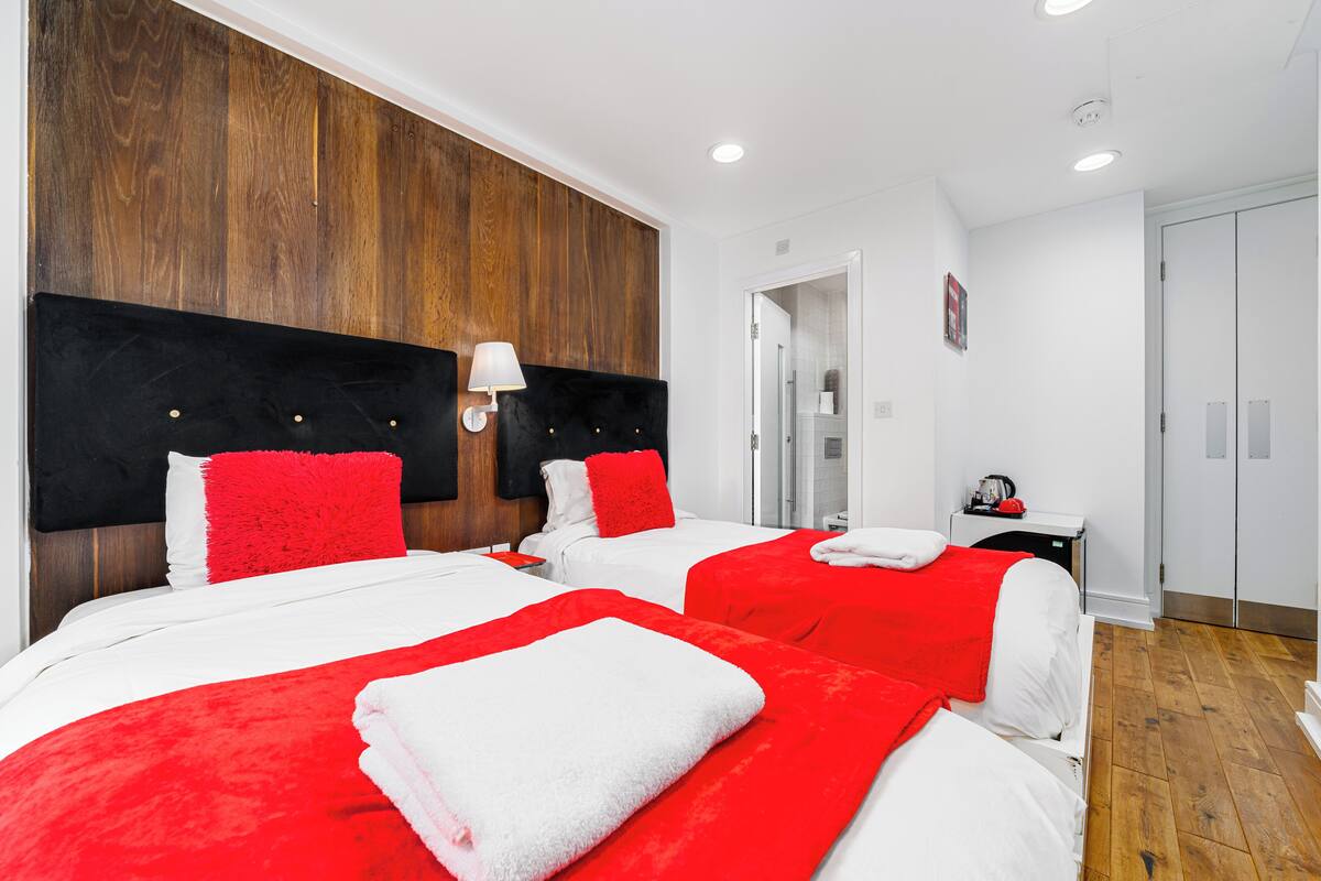 A hotel room with two beds, each with red and white bedding, a wooden accent wall, and a small bedside table with a lamp.
