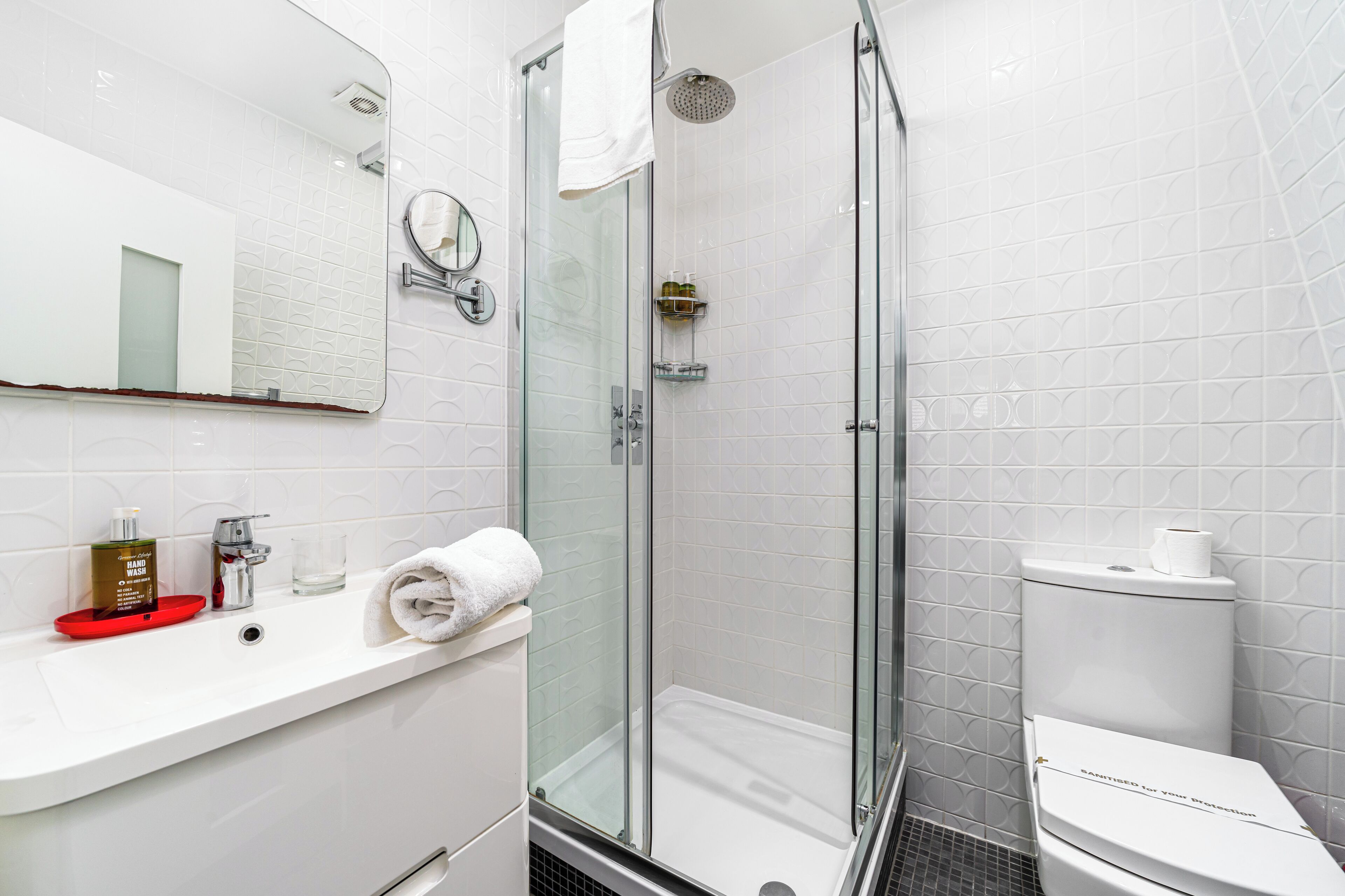 A modern bathroom with a glass-enclosed shower, a white sink with a red soap dish, and a toilet with a paper towel holder.