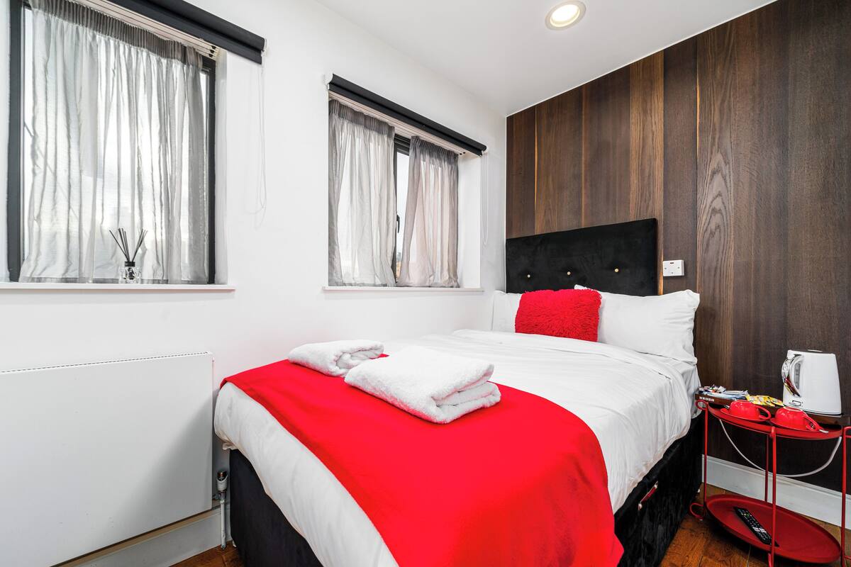 A modern hotel room with a large bed, red accents, a wooden accent wall, and a small side table with a kettle.