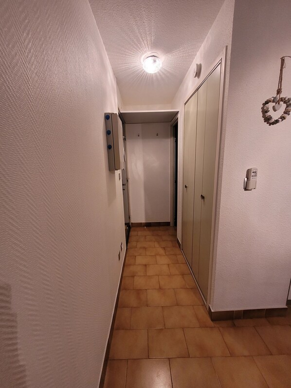 A narrow hallway with a wooden floor, white walls, and a ceiling light. There is a door at the end of the hallway and a wall-mounted light switch.