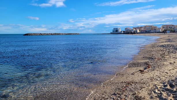 A beach with clear water, a rocky breakwater, and buildings in the background.